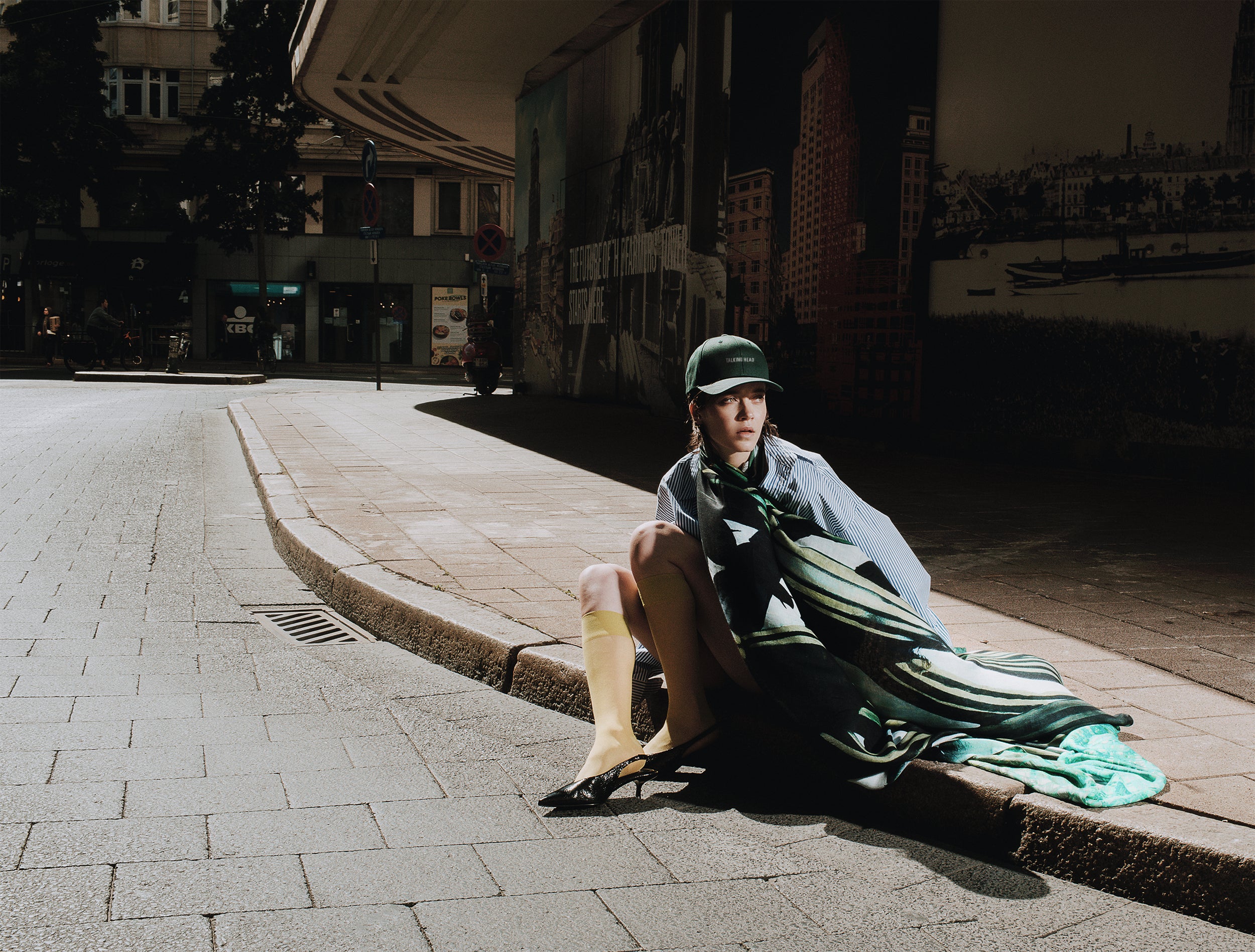 Person sitting on a city street wearing a green dress and hat.